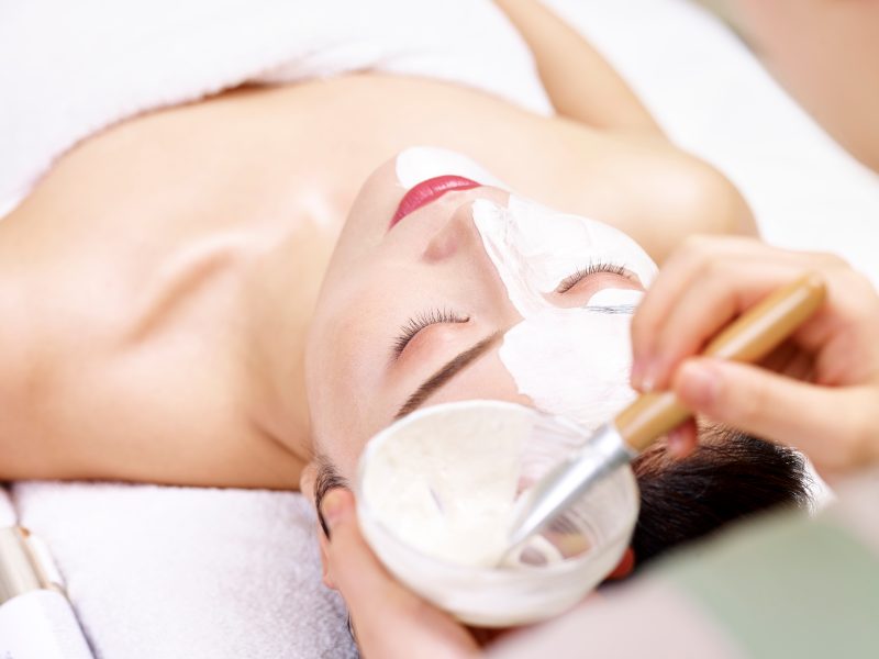 asian beautician applying facial mask on face of young woman